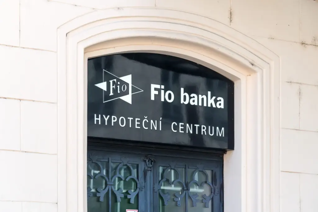 Vnější pohled na pobočku Fio banky v České republice. Budova se vyznačuje klasickým evropským architektonickým stylem s velkými prosklenými okny. Nad vchodem je výrazný bílozelený nápis s logem „Fio banka“. Fasáda je čistá a moderní, integrovaná do tradiční městské uliční krajiny.