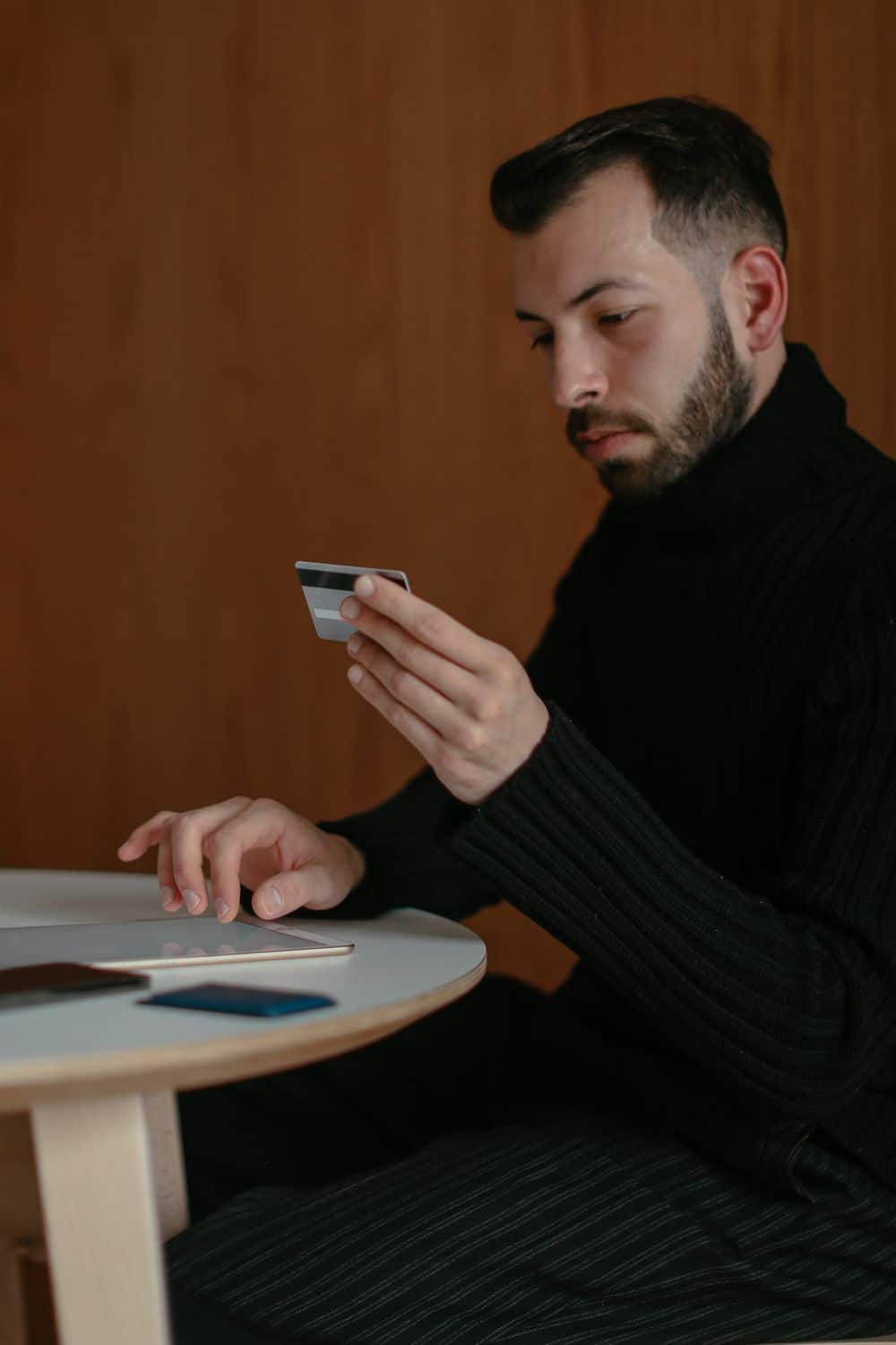 A young man sitting indoors, holding a credit card to make an online purchase.
