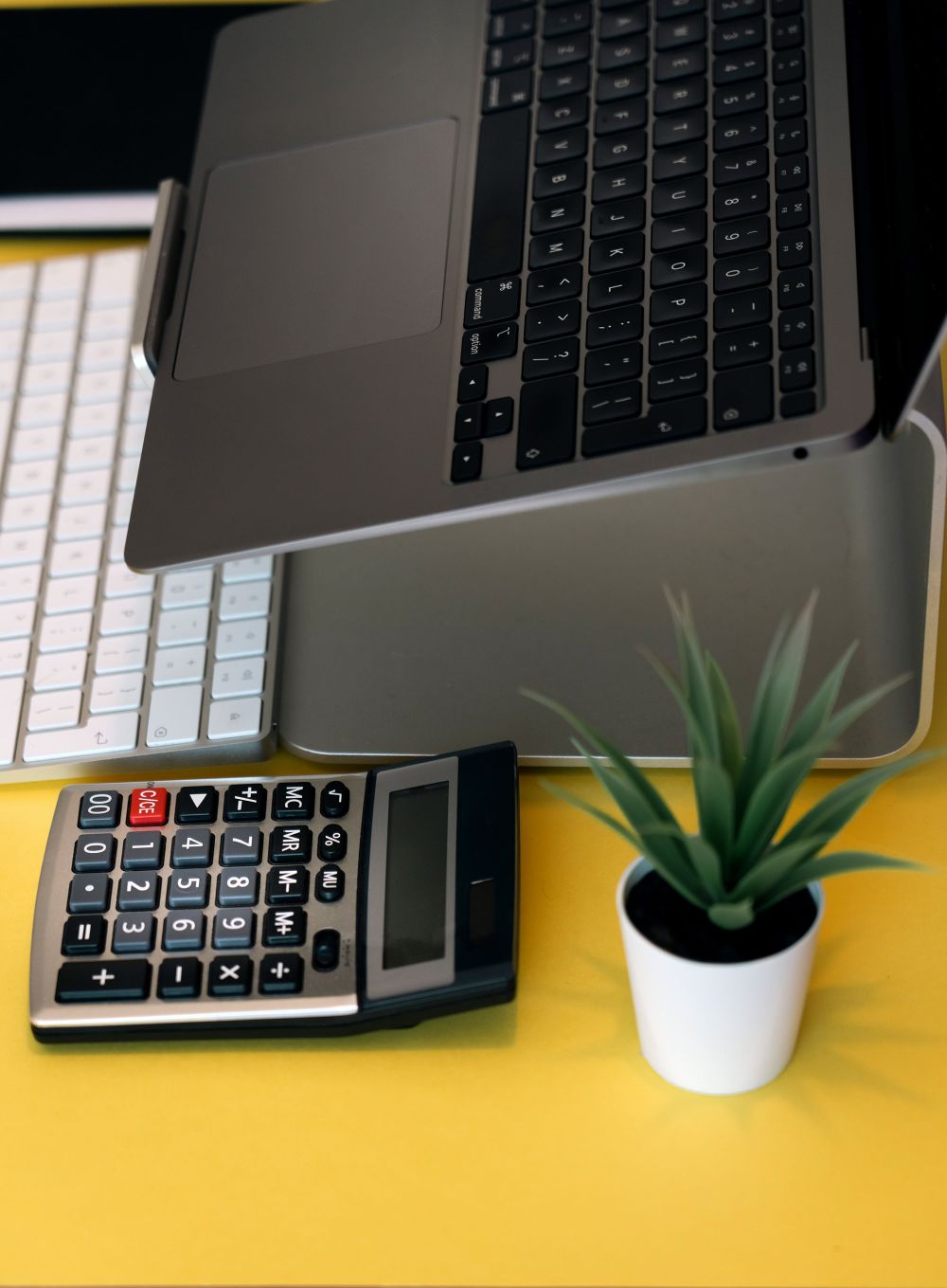 Vertical shot of a workspace with laptop, calculator, and potted plant on a vibrant yellow desk.
