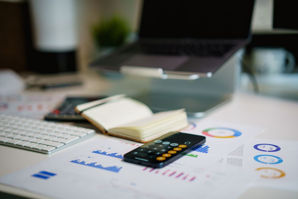 Modern office desk with a laptop, smartphone calculator app, and financial charts.