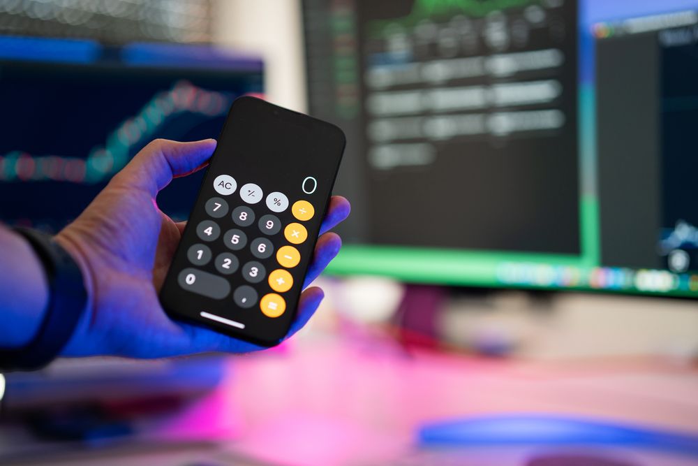 A person uses a smartphone calculator app in a modern office with computer screens displaying charts.