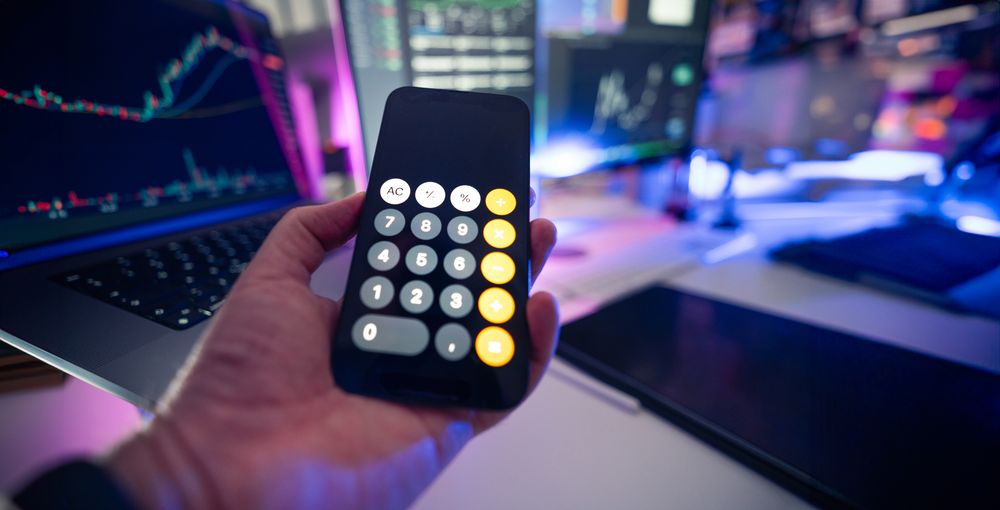 Smartphone with calculator app against financial charts on monitors, symbolizing financial planning.