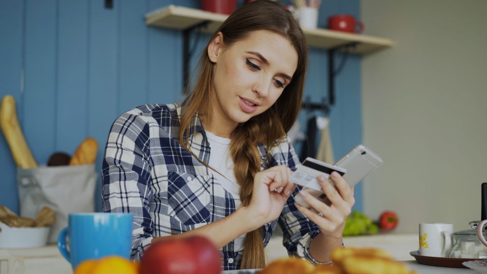 Woman in plaid shirt using credit card with smartphone in cozy kitchen setting.