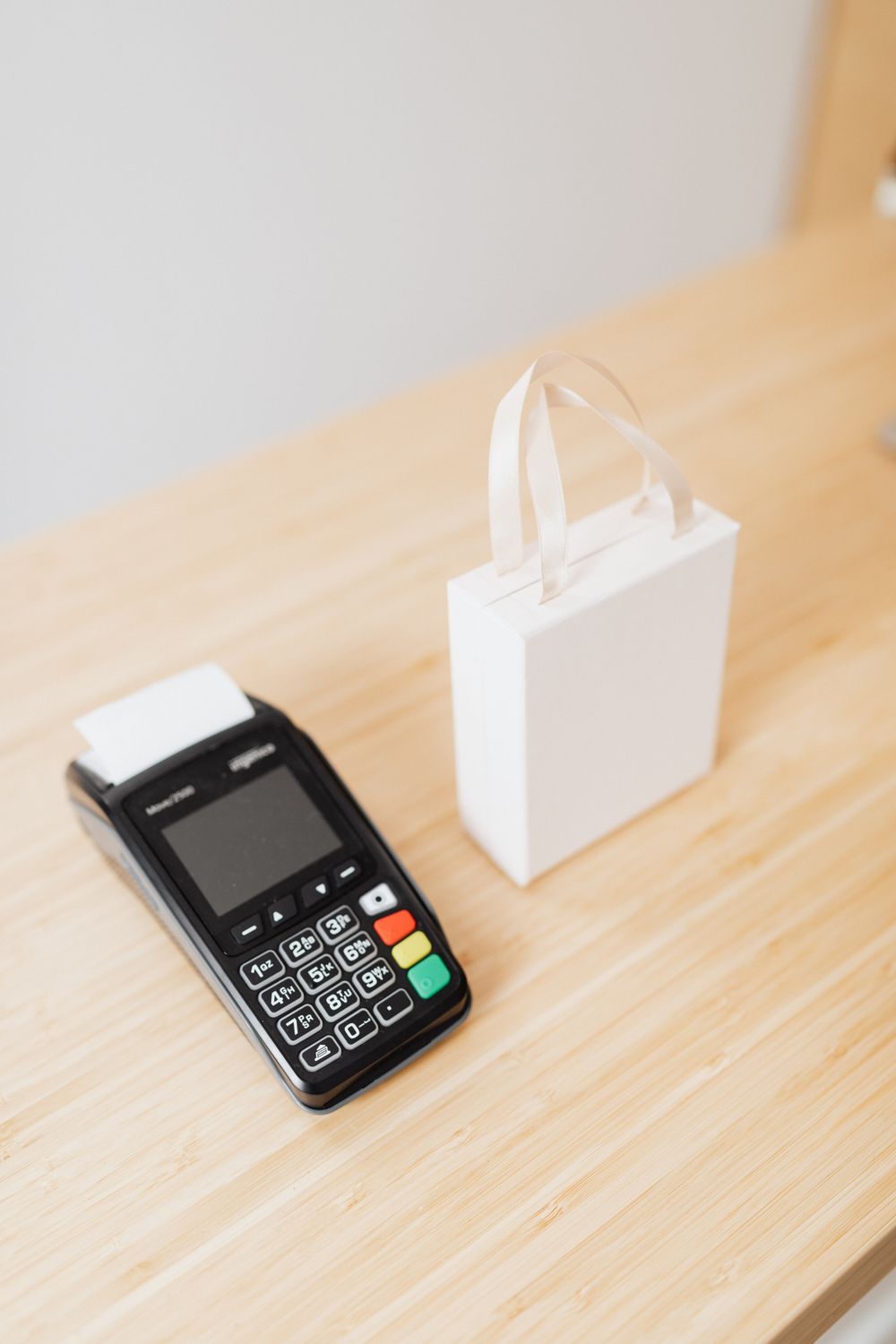 A payment terminal and a small white gift bag on a wooden countertop, ideal for retail or shopping themes.