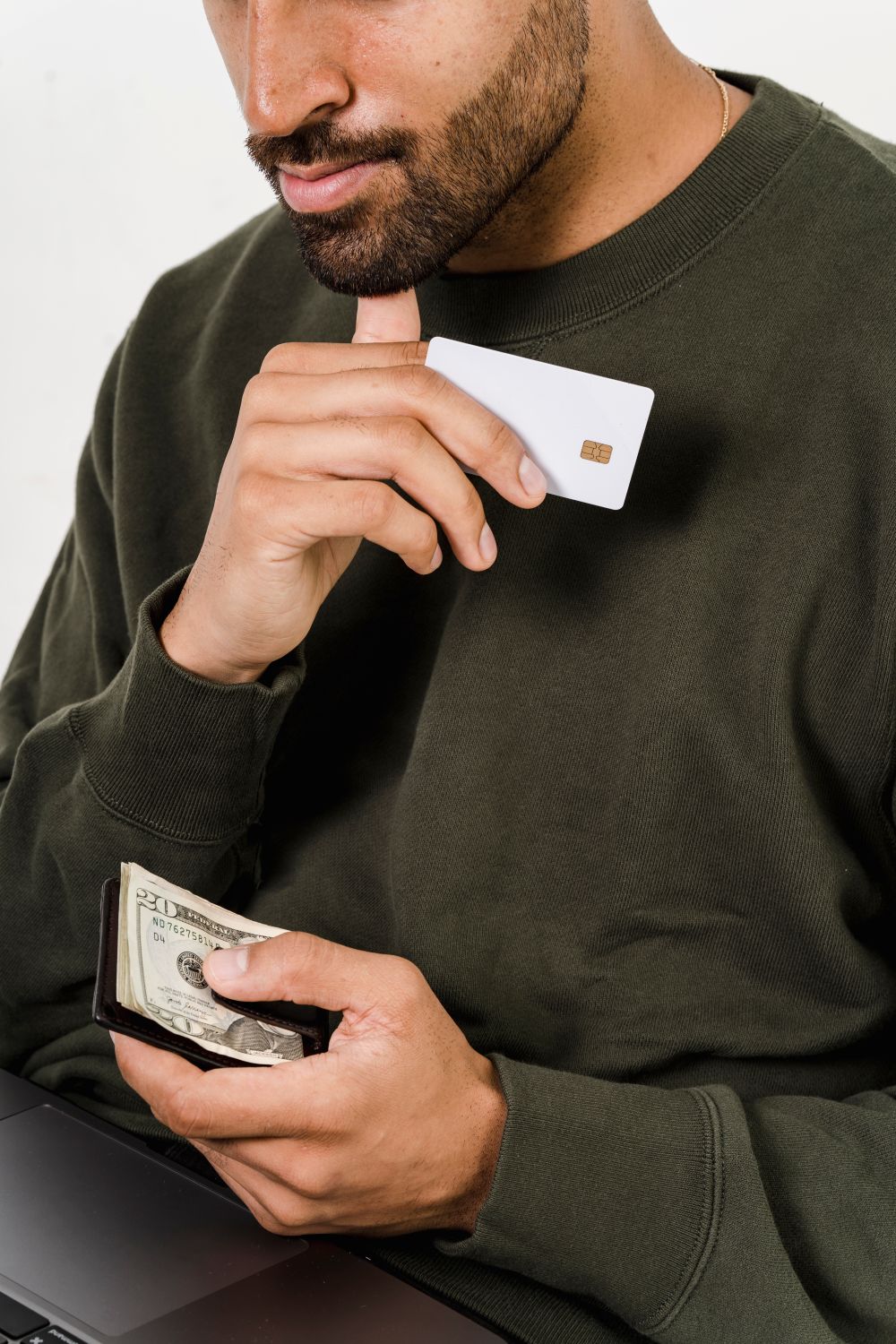 A man in a green sweater holds a credit card and cash, contemplating his payment options.