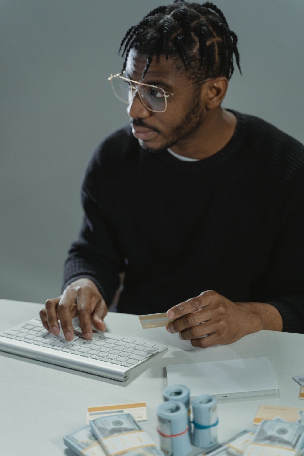 Man with glasses using a keyboard, holding a bank card, surrounded by cash, suggesting online finance or cybercrime themes.