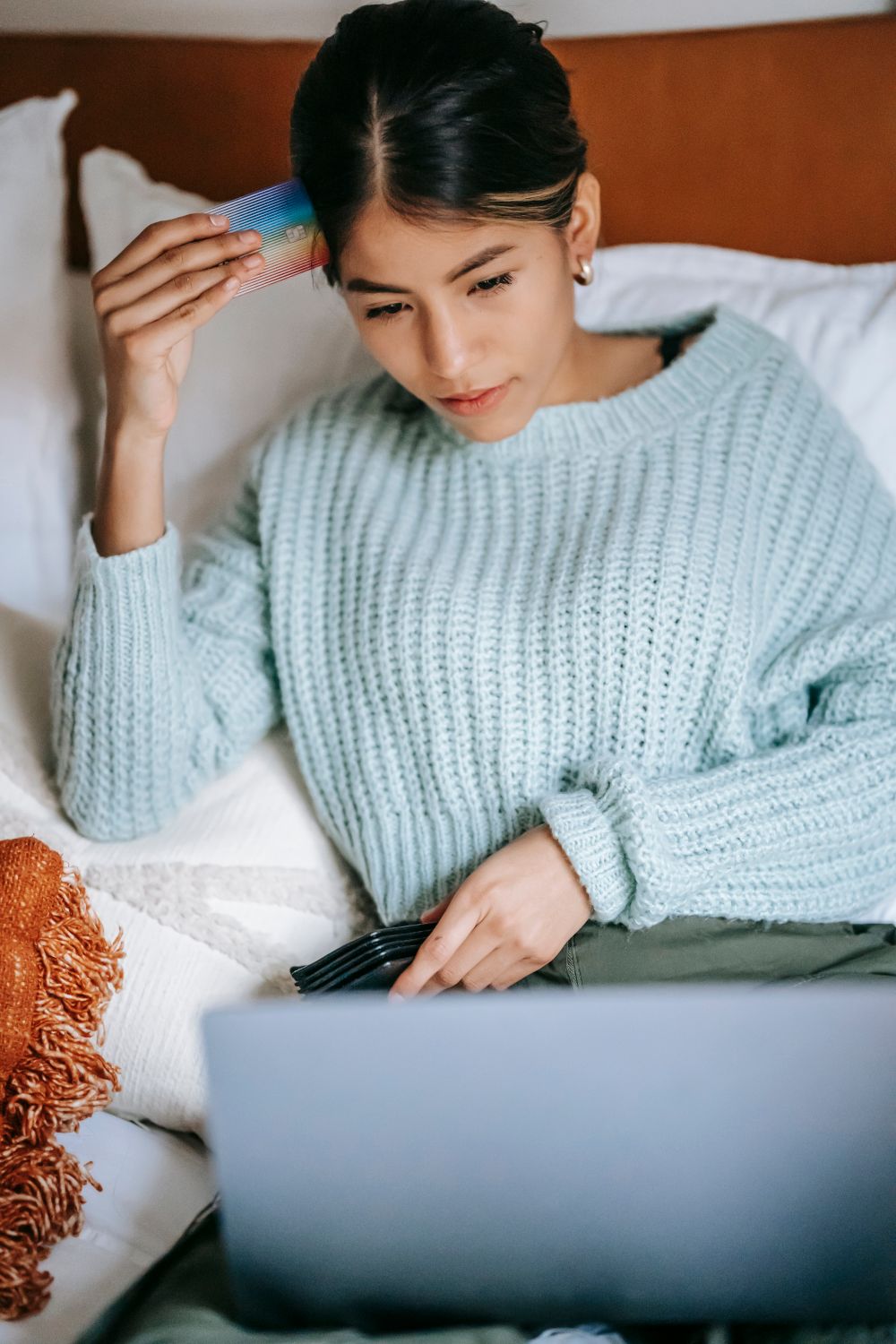Focused young Asian female in blue sweater making online purchase via netbook while lying on bed with credit card