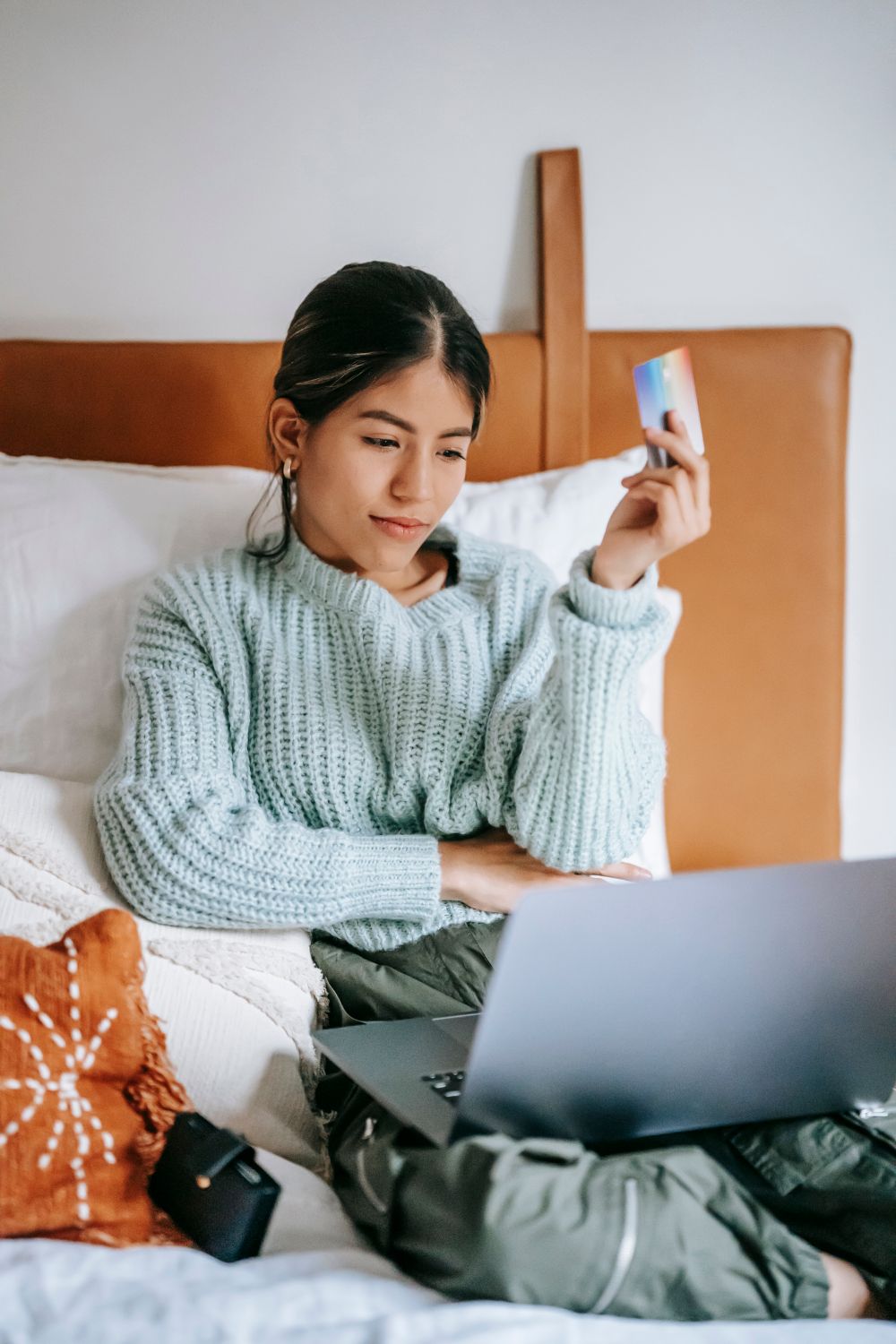 Calm young Asian female in casual wear making online payment via netbook while lying on comfortable bed with credit card