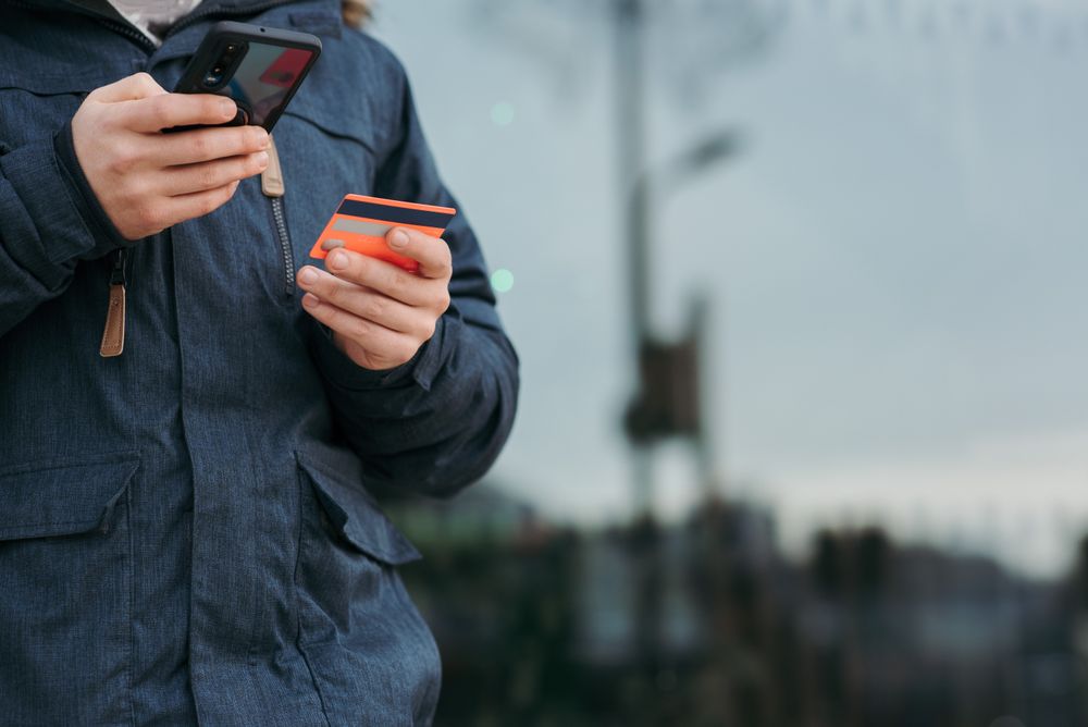 Crop anonymous man in warm clothes holding credit card and browsing smartphone while standing on street in daytime