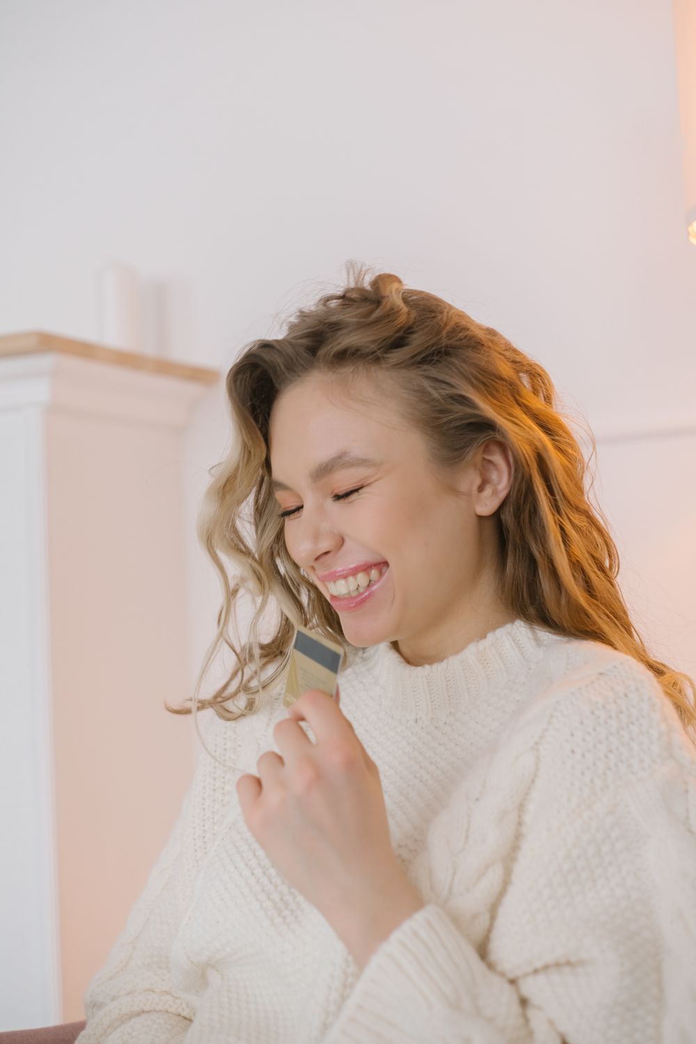 Happy young woman in a cozy sweater holding a credit card indoors, expressing joy and contentment.
