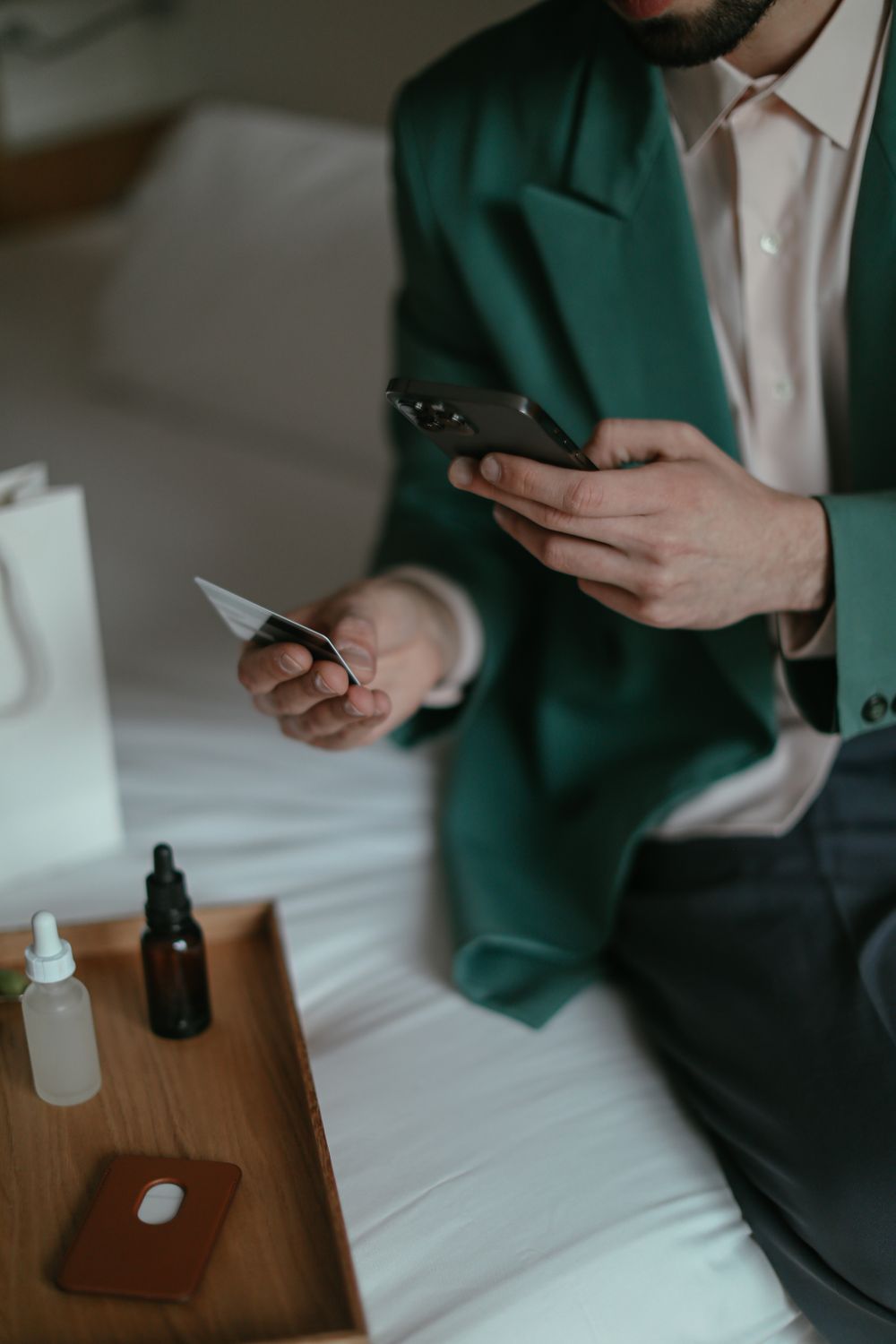 Man holding a credit card and smartphone for online shopping, sitting on a bed with beauty products nearby.