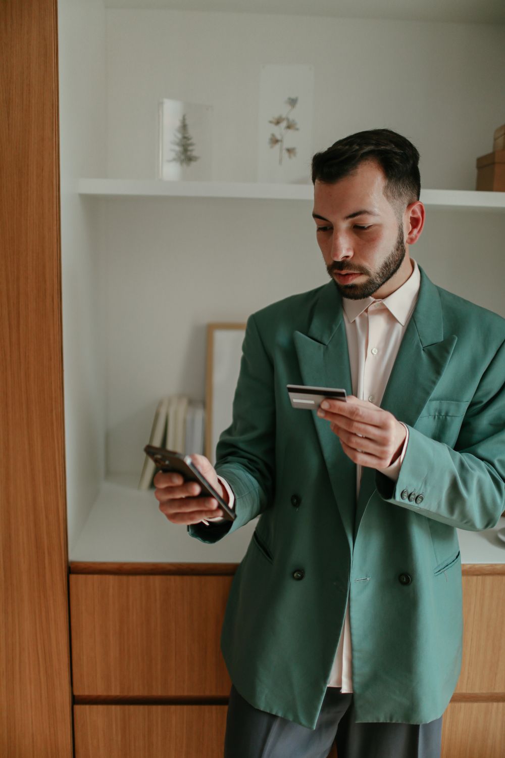 Businessman using smartphone and credit card with wooden bookshelf in background. Indoor setting.