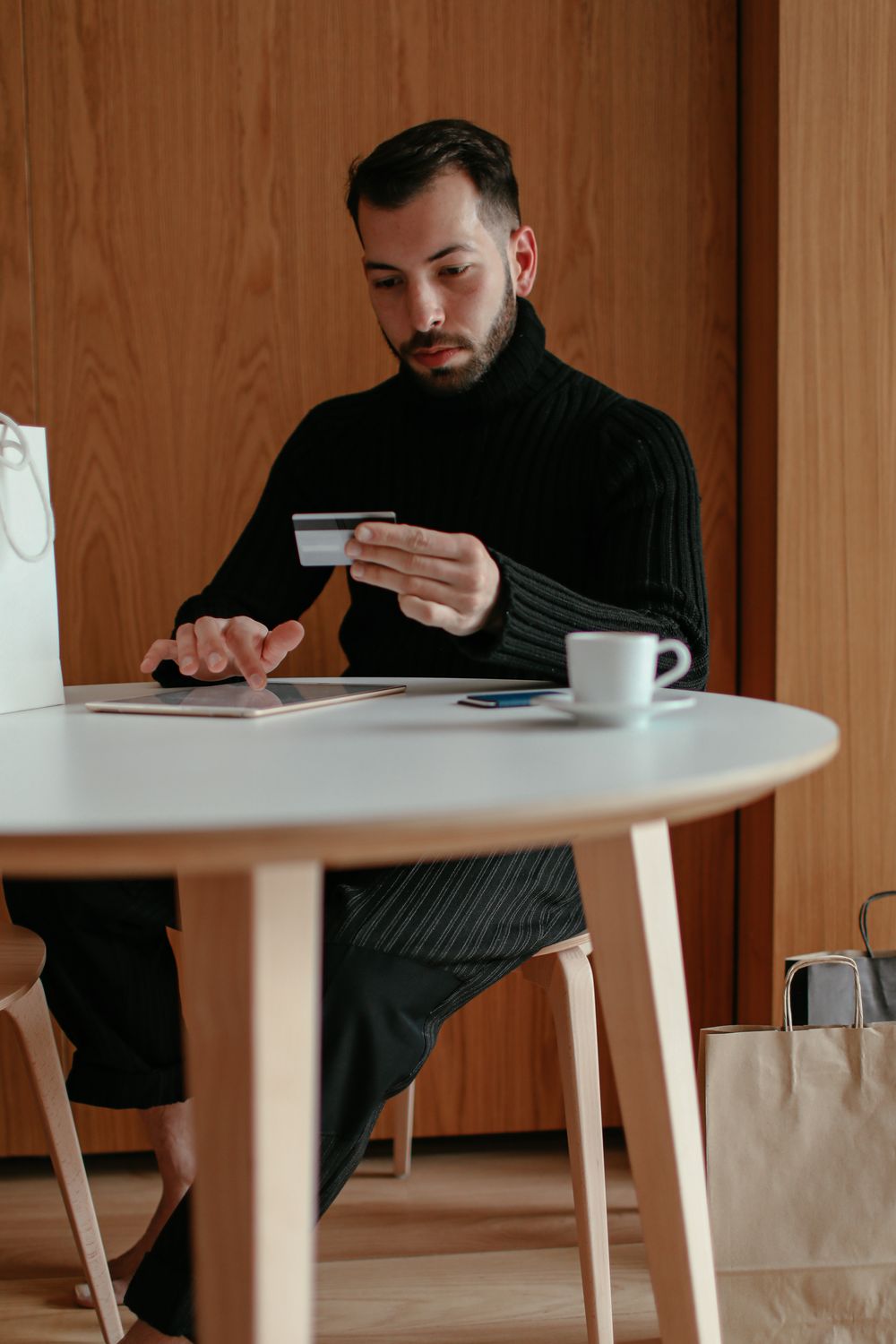 Concentrated young bearded male sitting at table with cup of coffee and browsing tablet while doing online purchase with credit card
