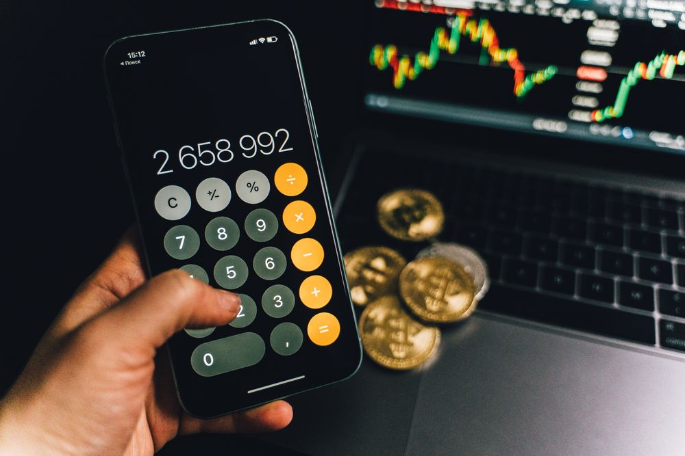 A smartphone displays a calculator app beside bitcoins and a laptop with a financial chart.
