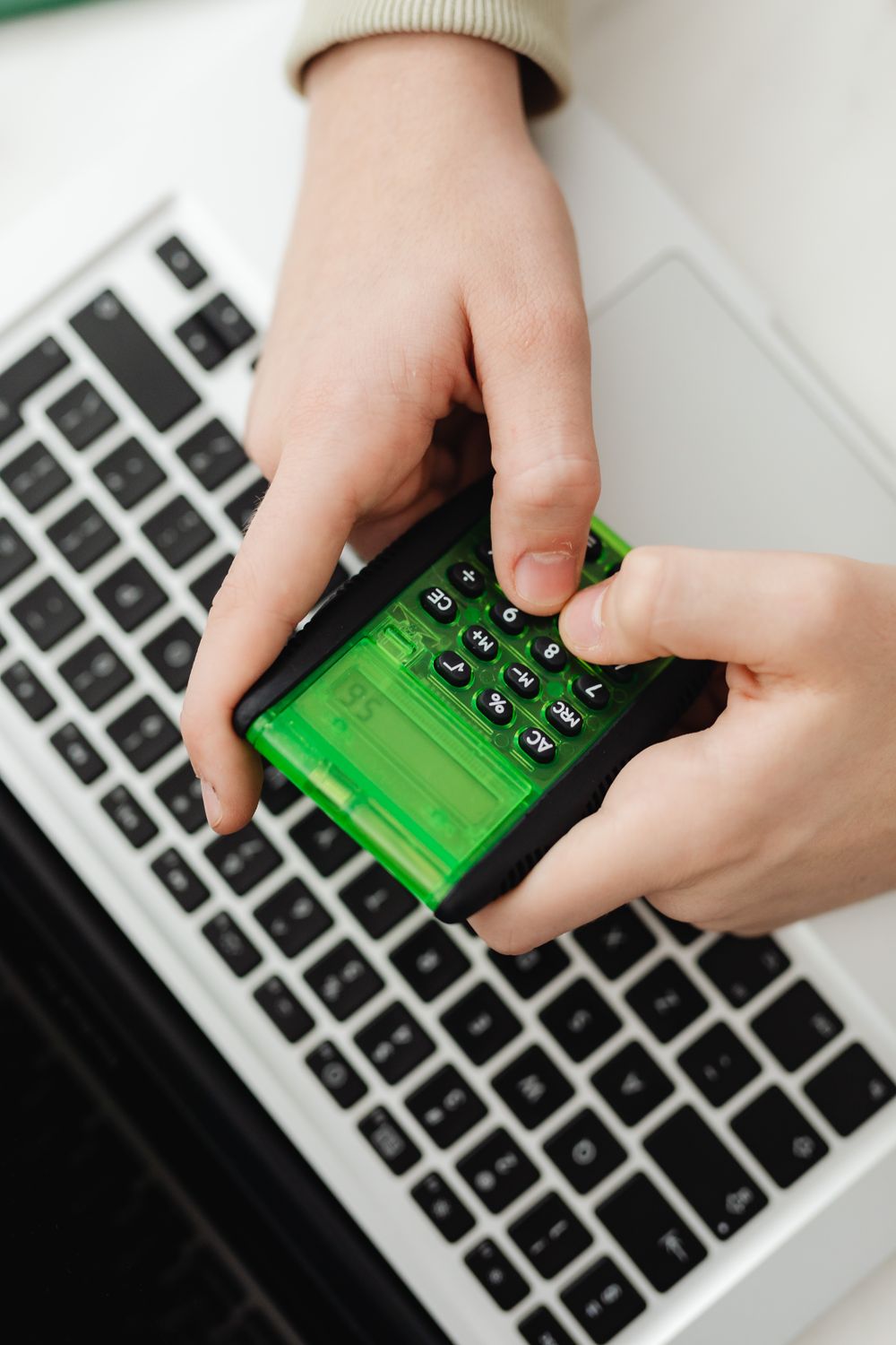 Hands operating a green calculator over a laptop keyboard in an indoor setting.
