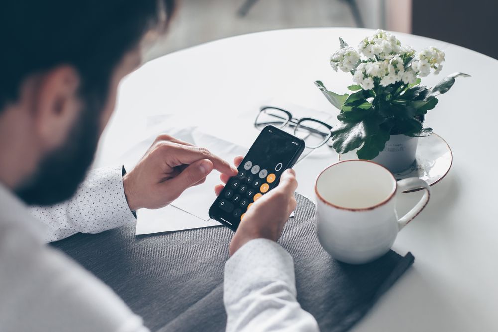 Adult man using smartphone calculator at table with flowers and coffee cup.