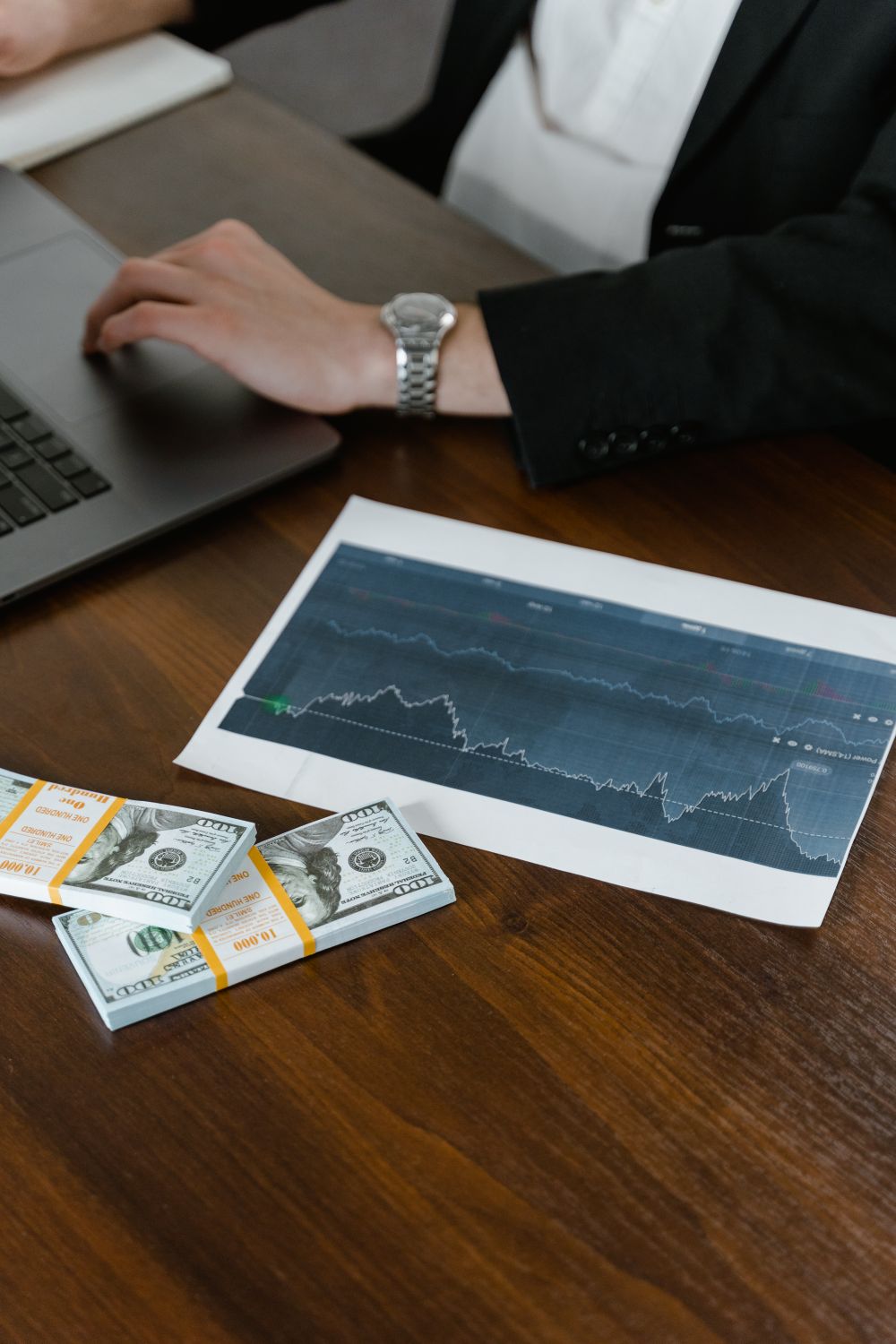 A desk scene with cash bundles, a chart printout, and a person working on a laptop.