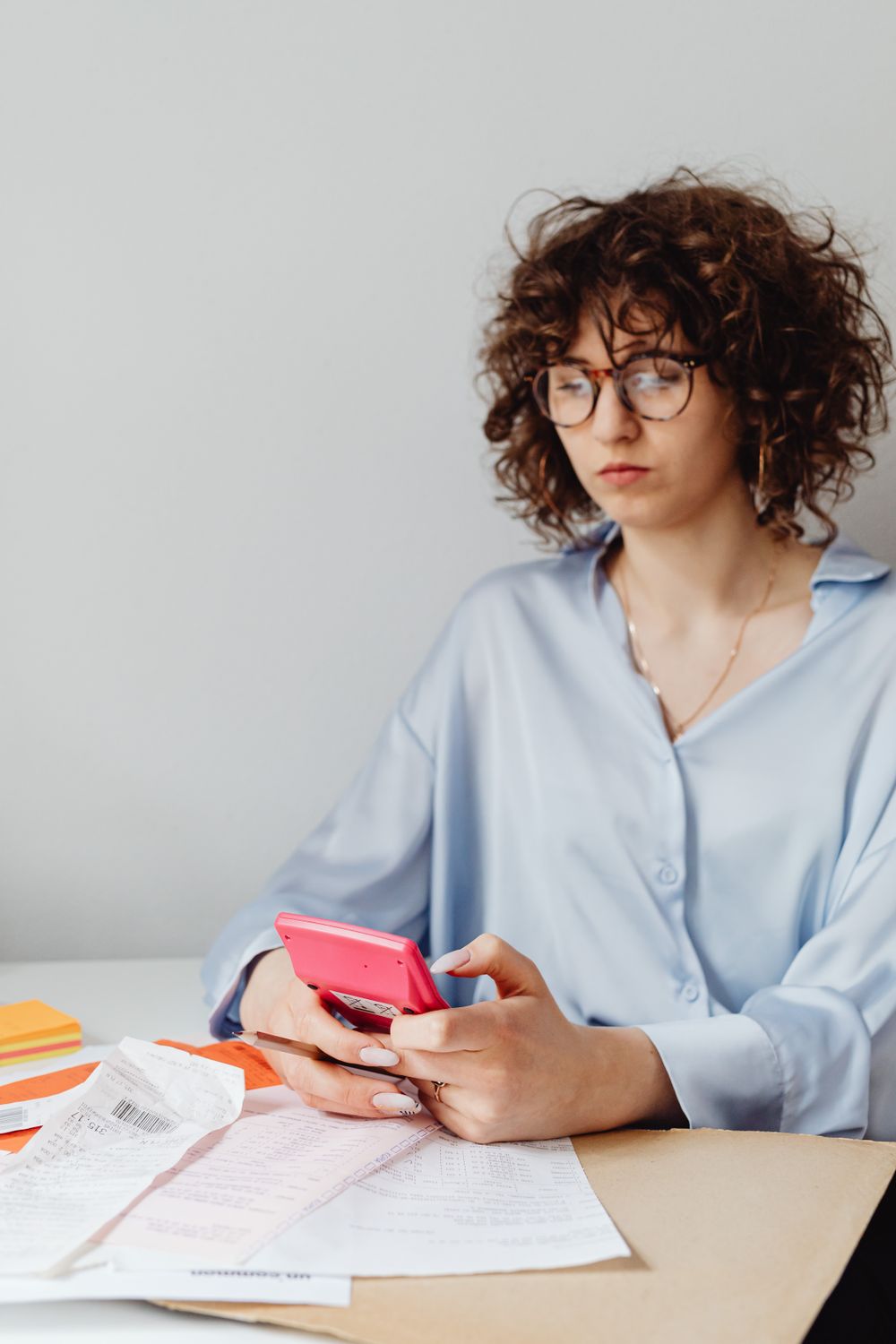 Woman with curly hair and eyeglasses reviewing bills and using a calculator at a desk indoors.